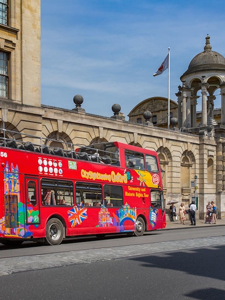 Red double-decker bus on Oxford street near historic building, part of Oxford by Rail day tour from London.
