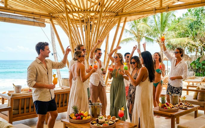 Group enjoying drinks at FINNS Beach Party Lagoon Zone, Bali, under bamboo structure by the ocean.