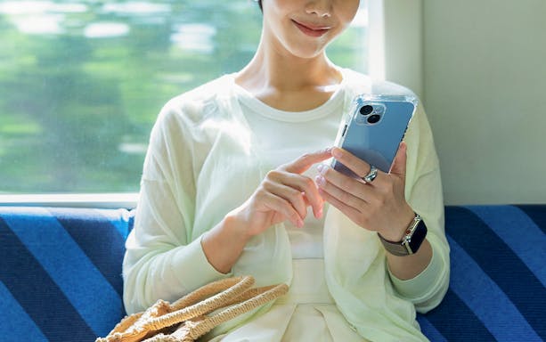 Woman on train using cellphone, seated by window with woven bag.
