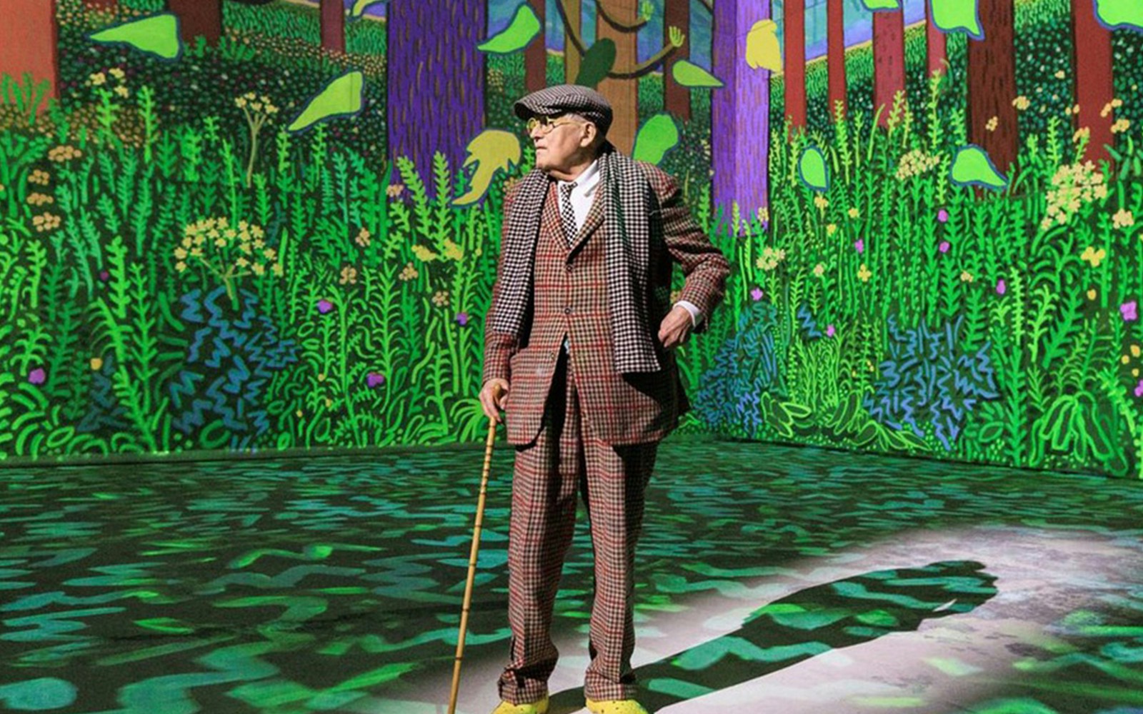 Man in checkered suit with cane in front of vibrant forest artwork, David Hockney: Bigger & Closer exhibit.