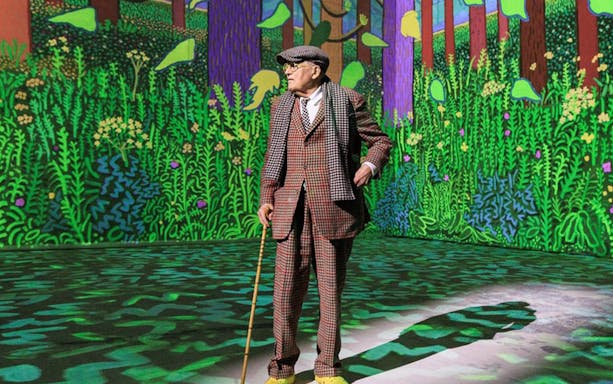Man in checkered suit with cane in front of vibrant forest artwork, David Hockney: Bigger & Closer exhibit.