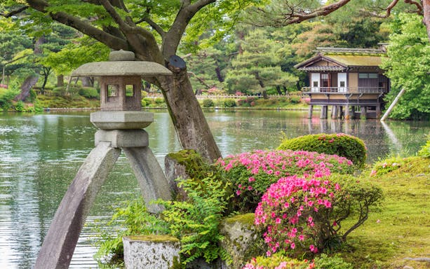 Stone lantern by pond in Kenrokuen Garden, Kanazawa, Japan, with traditional teahouse in background.