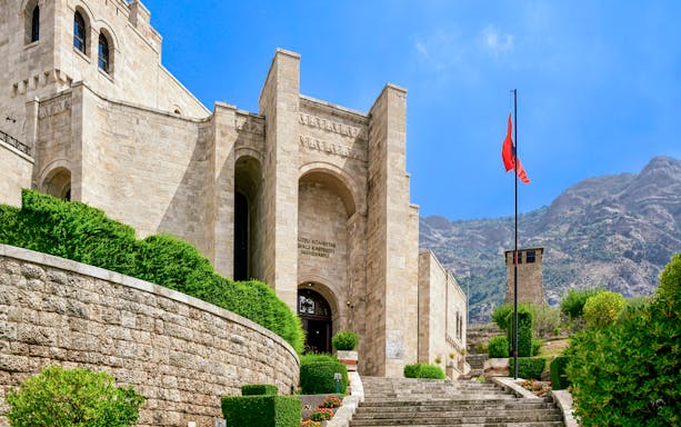 Skanderbeg Museum entrance at Kruje Castle, Albania, with Albanian flag and mountain backdrop.