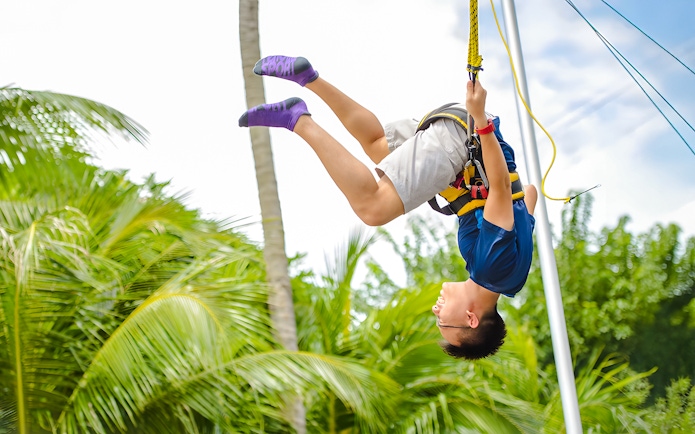 Person ziplining at Mega Adventure Park, Singapore, surrounded by palm trees.