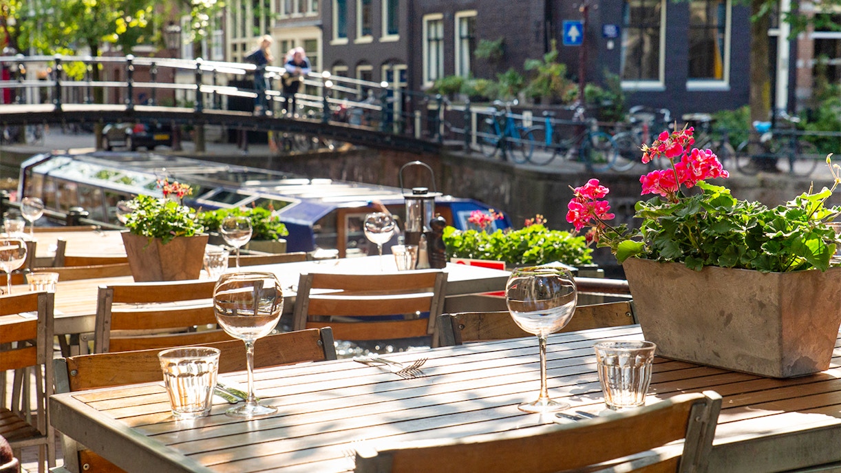 Outdoor cafe table by canal in Amsterdam's Jordaan District with flowers and bridge.
