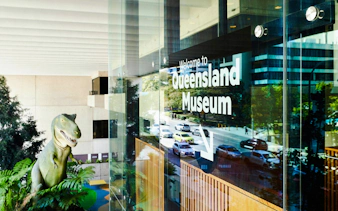 Queensland Museum entrance with dinosaur statue visible through glass.