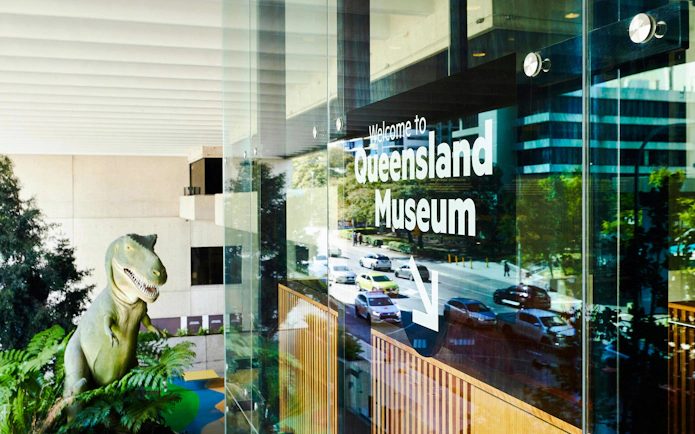Queensland Museum entrance with dinosaur statue visible through glass.