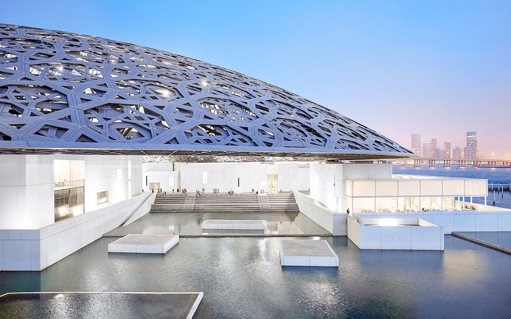 Louvre Abu Dhabi's dome and water features at sunset with city skyline in background.