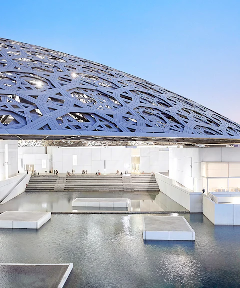 Louvre Abu Dhabi's dome and water features at sunset with city skyline in background.