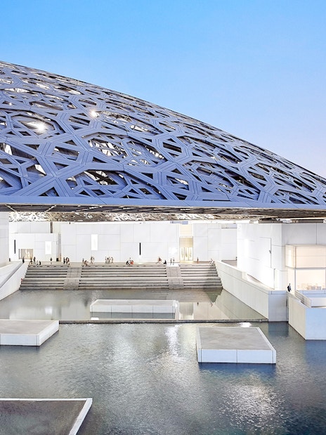 Louvre Abu Dhabi's dome and water features at sunset with city skyline in background.