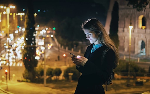 Visitor using phone near illuminated Roman Forum at night, Rome, Italy.