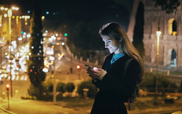 Visitor using phone near illuminated Roman Forum at night, Rome, Italy.