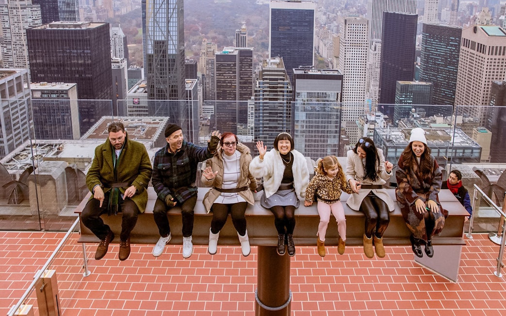 People sitting on The Beam at Top of the Rock, Rockefeller Center, New York City skyline in background.