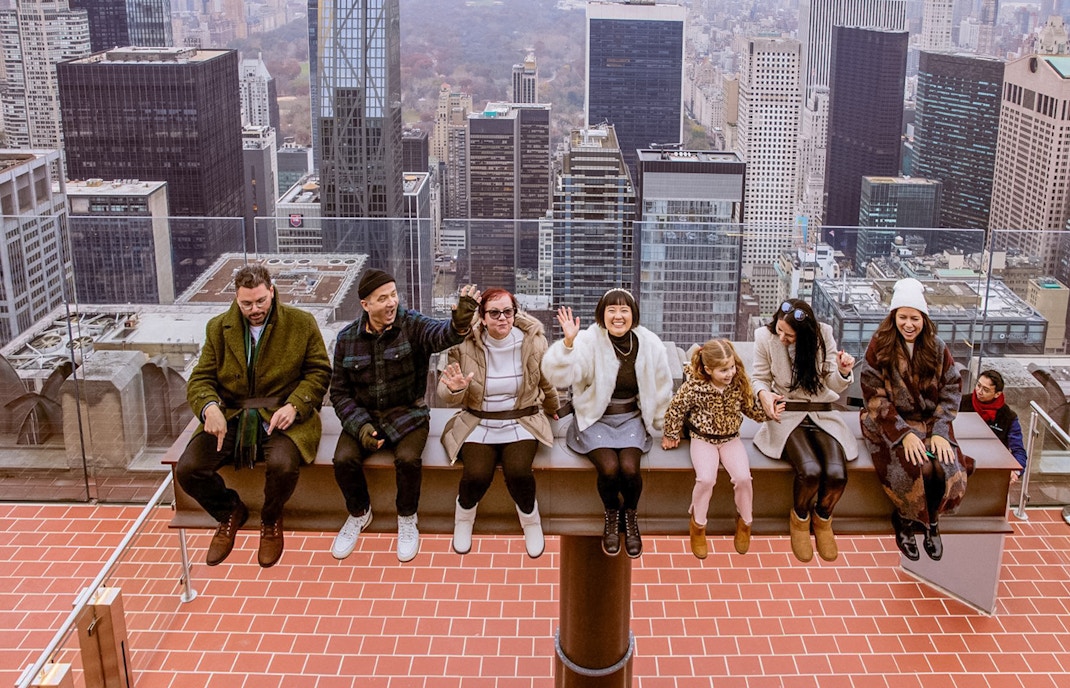 People sitting on The Beam at Top of the Rock, Rockefeller Center, New York City skyline in background.