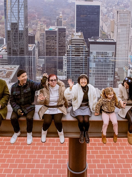 People sitting on The Beam at Top of the Rock, Rockefeller Center, New York City skyline in background.