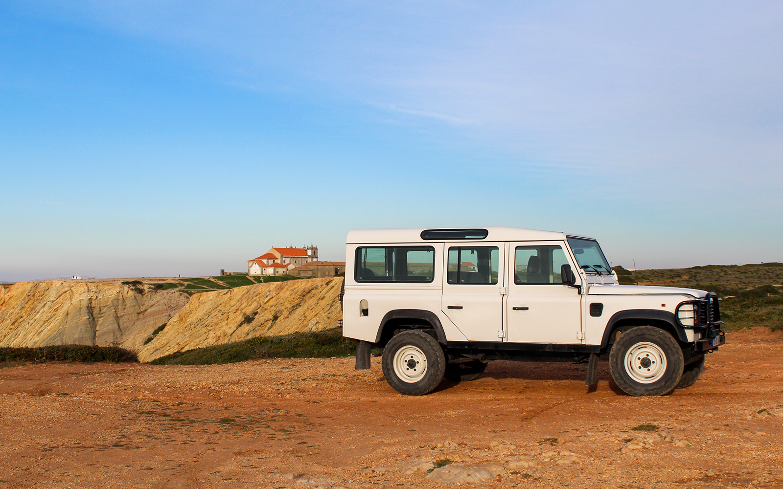 4x4 Jeep parked near Cape Espichel cliffs with a view of a historic building in the background.