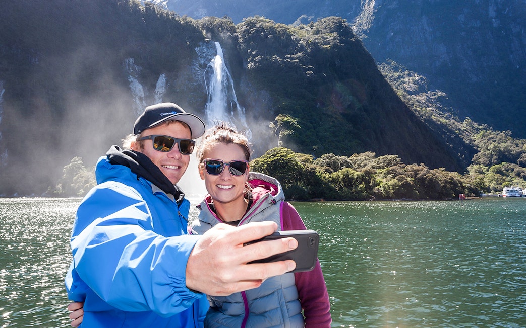 Couple taking selfie on Milford Sound cruise with waterfall in background.