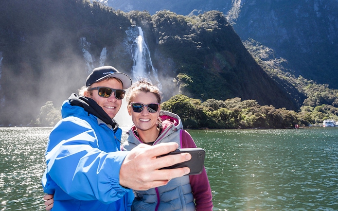 Couple taking selfie on Milford Sound cruise with waterfall in background.
