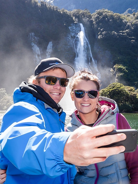 Couple taking selfie on Milford Sound cruise with waterfall in background.