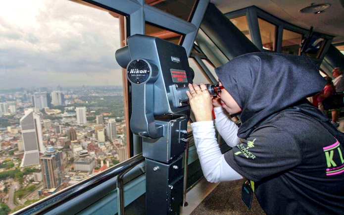 Visitor using binoculars at KL Tower observation deck, overlooking Kuala Lumpur cityscape.