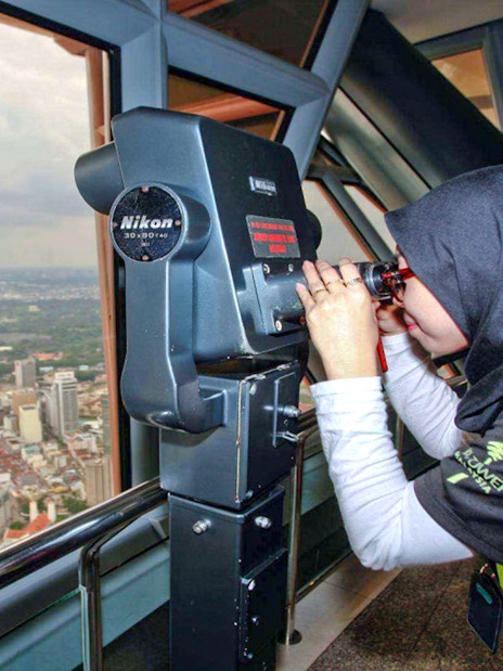 Visitor using binoculars at KL Tower observation deck, overlooking Kuala Lumpur cityscape.