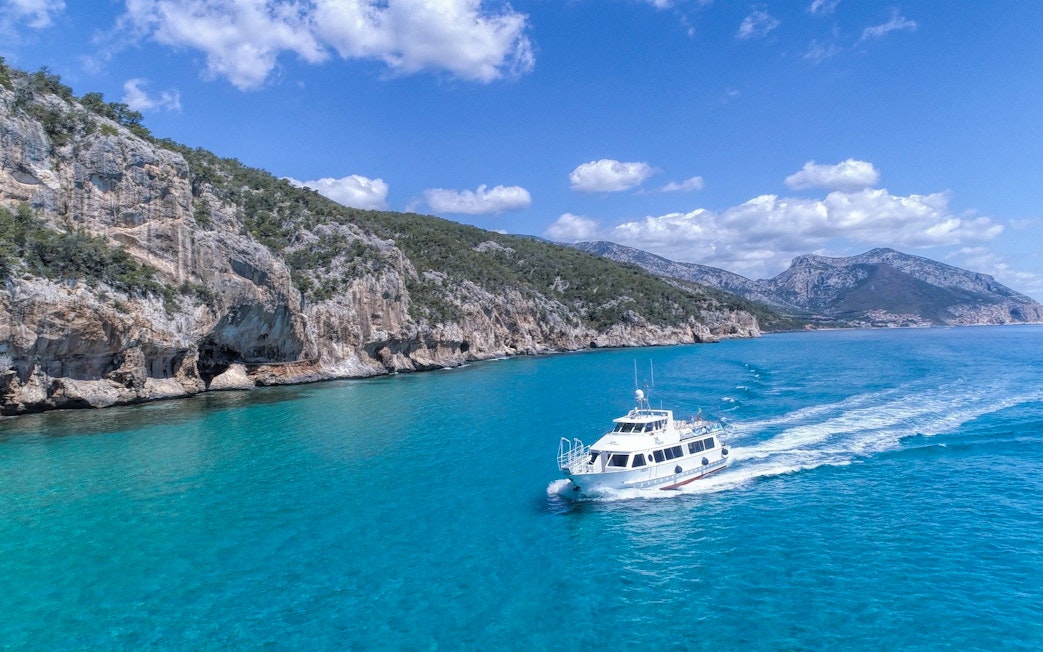 Boat cruising in the turquoise waters of Gulf of Orosei, Italy with rocky cliffs in the background.