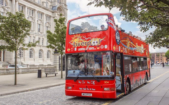 Red double-decker bus on Liverpool Hop On Hop Off City Tour near historic building.