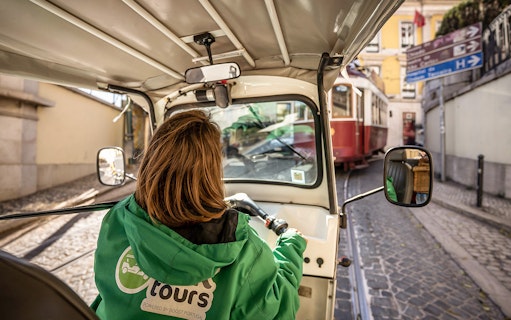 Person driving an electric tuk tuk on a cobblestone street in Lisbon, passing a red tram.