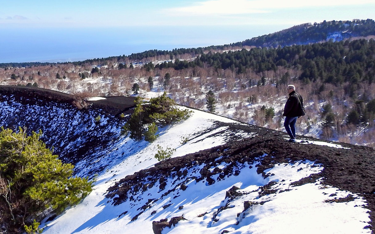 Person walking on snowy slope of Mount Etna with forested landscape in background.