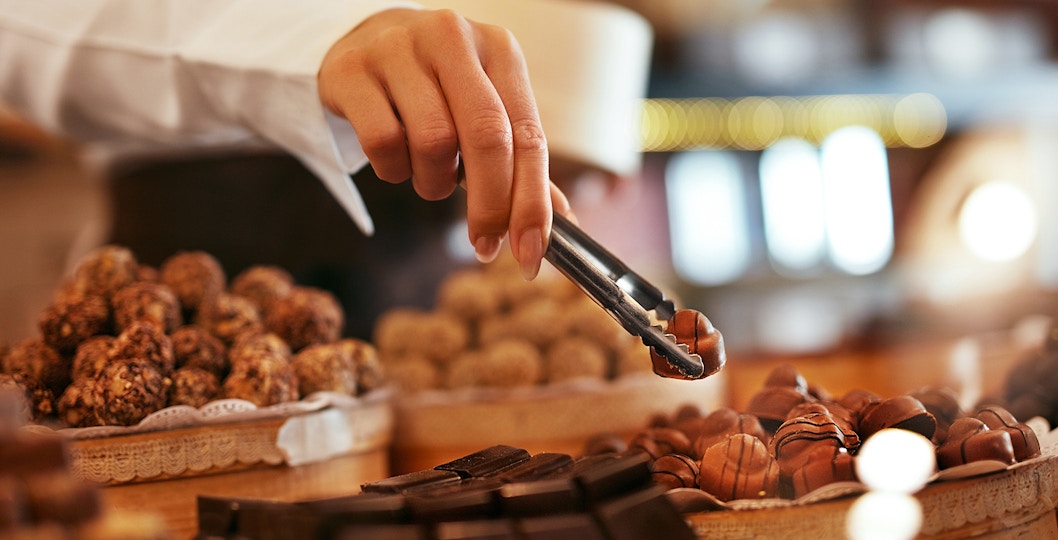 The image shows a person using tongs to pick up a chocolate from a display of various chocolates.