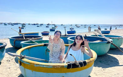 Women sitting in traditional round boats on Mui Ne beach, Vietnam, with fishing boats in the background.