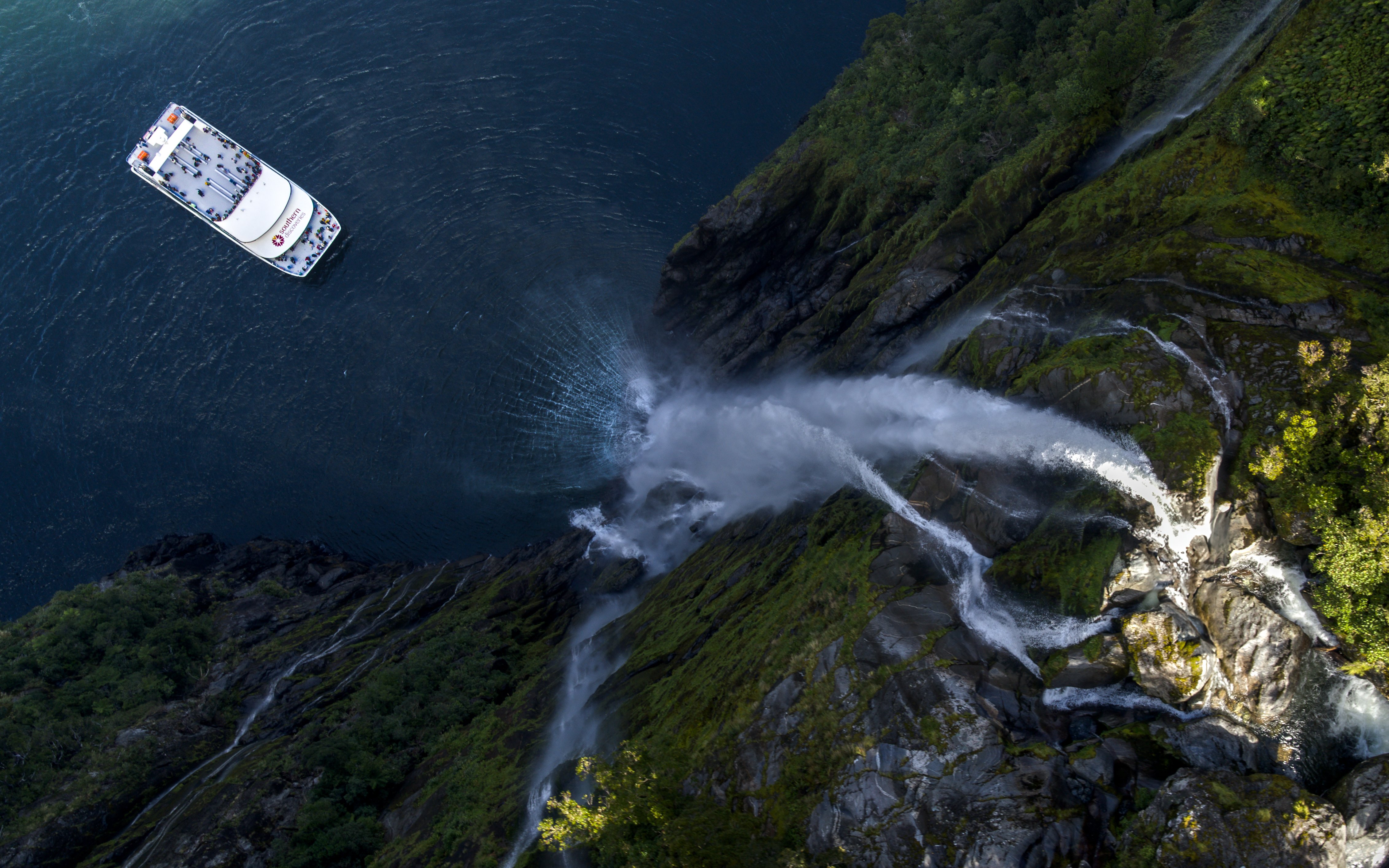 Cruise boat near waterfall in Milford Sound, New Zealand, surrounded by lush cliffs.