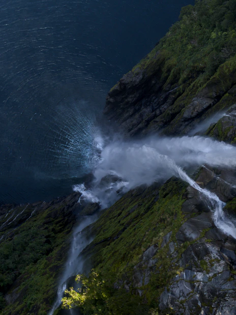 Cruise boat near waterfall in Milford Sound, New Zealand, surrounded by lush cliffs.