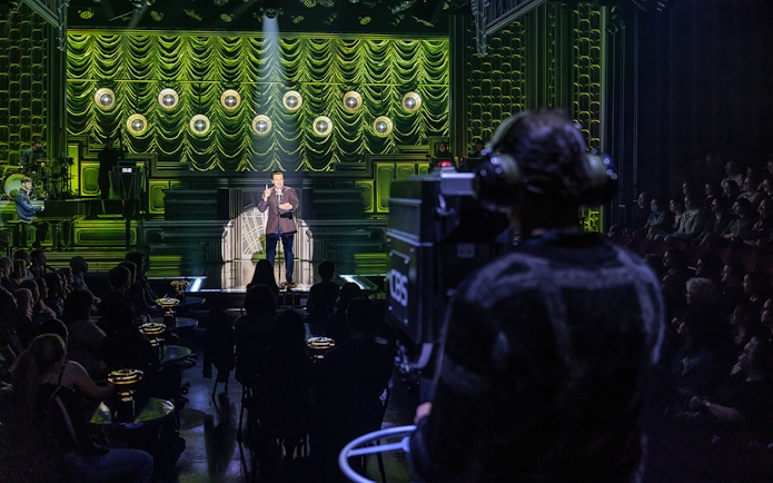 Performer on stage with audience and camera operator, theater setting, "Just In Time" show.
