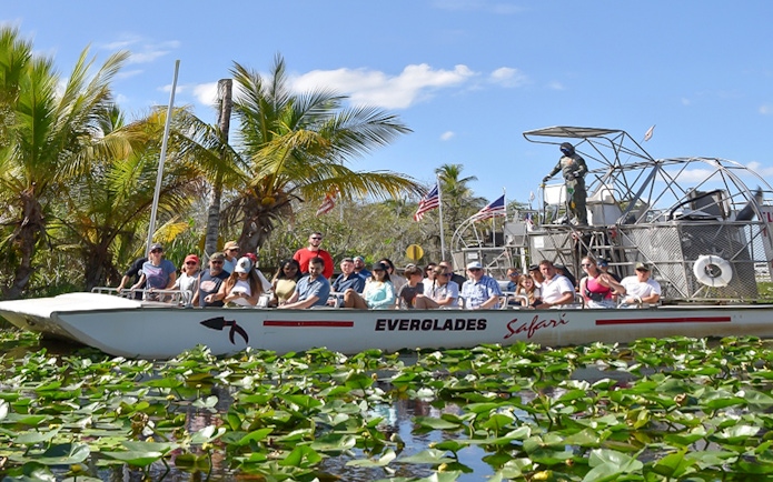 Tourists on an airboat ride through Everglades Safari Park, Miami, surrounded by palm trees and water lilies.