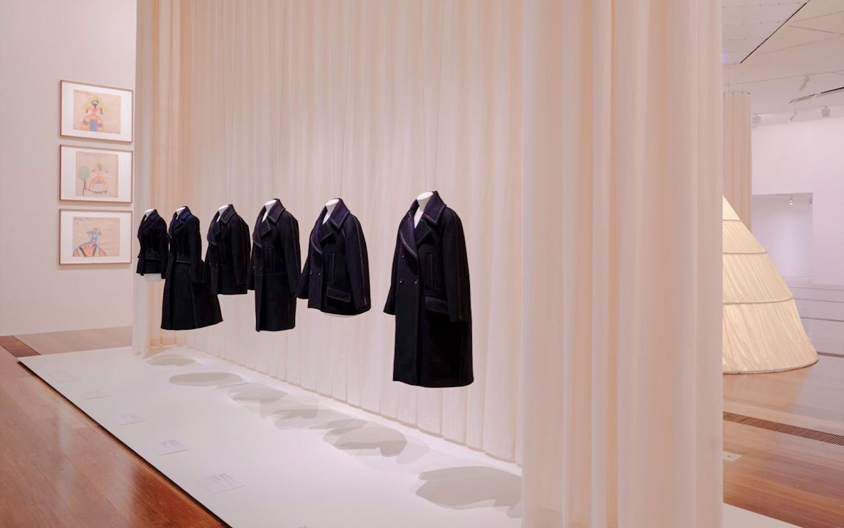 Coats displayed at Martin Grant Exhibition with framed artwork on the wall.