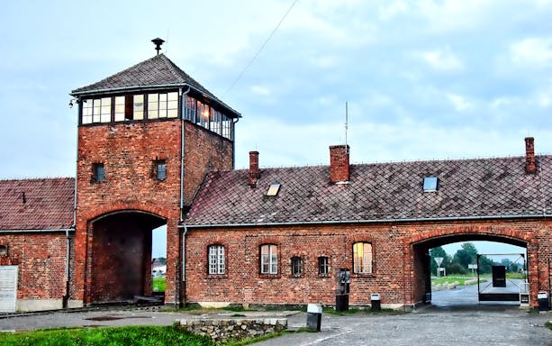 Auschwitz-Birkenau entrance gate with brick buildings under a cloudy sky.