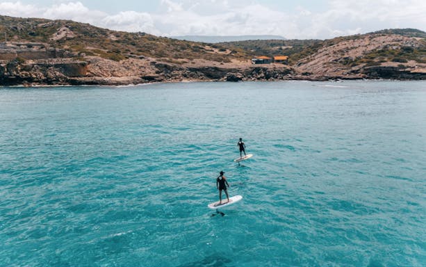 Two people e-surfing in the Gulf of Alghero with rocky coastline in the background.