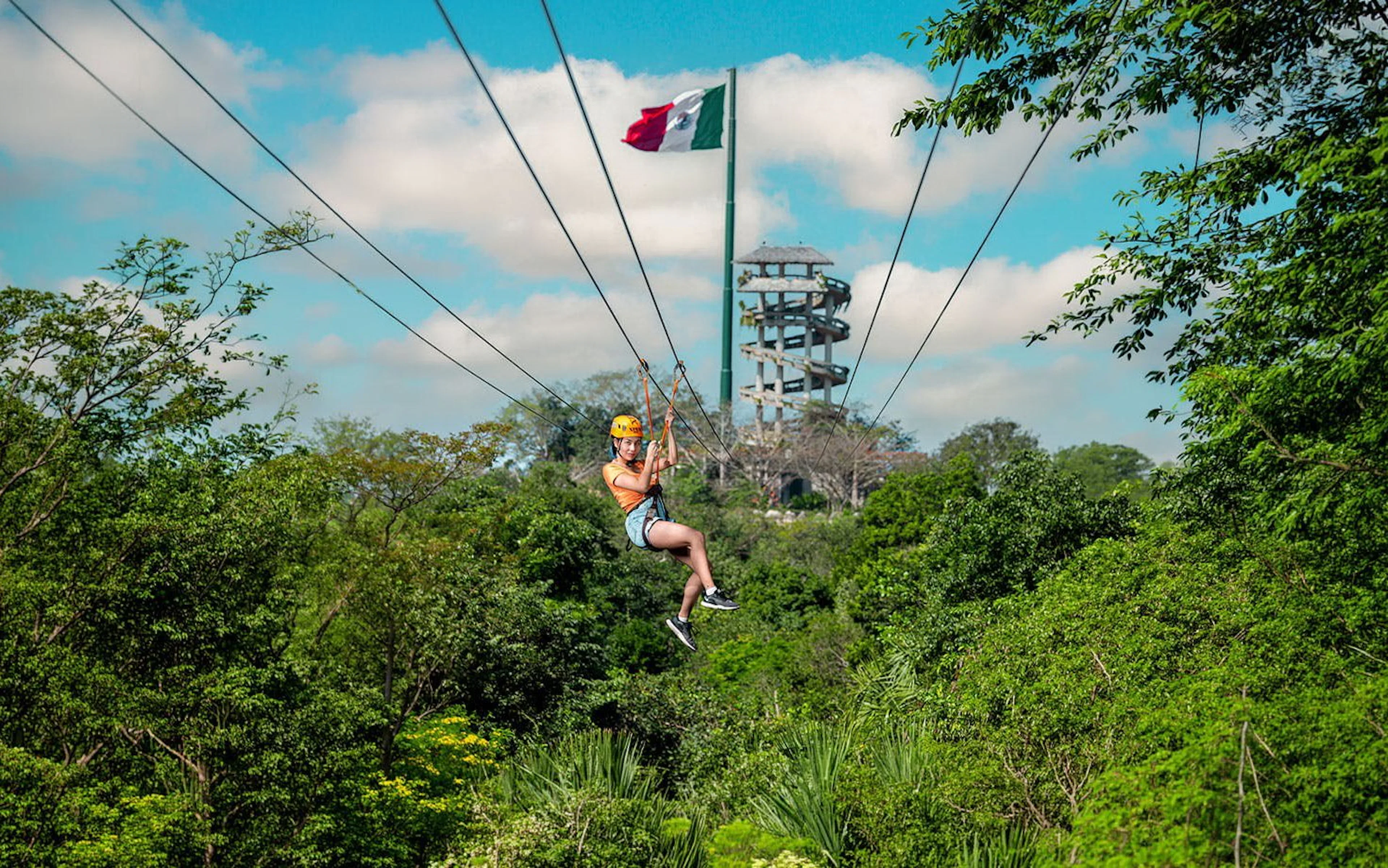 Ziplining over lush forest at Xplor Park with observation tower and flag in background.
