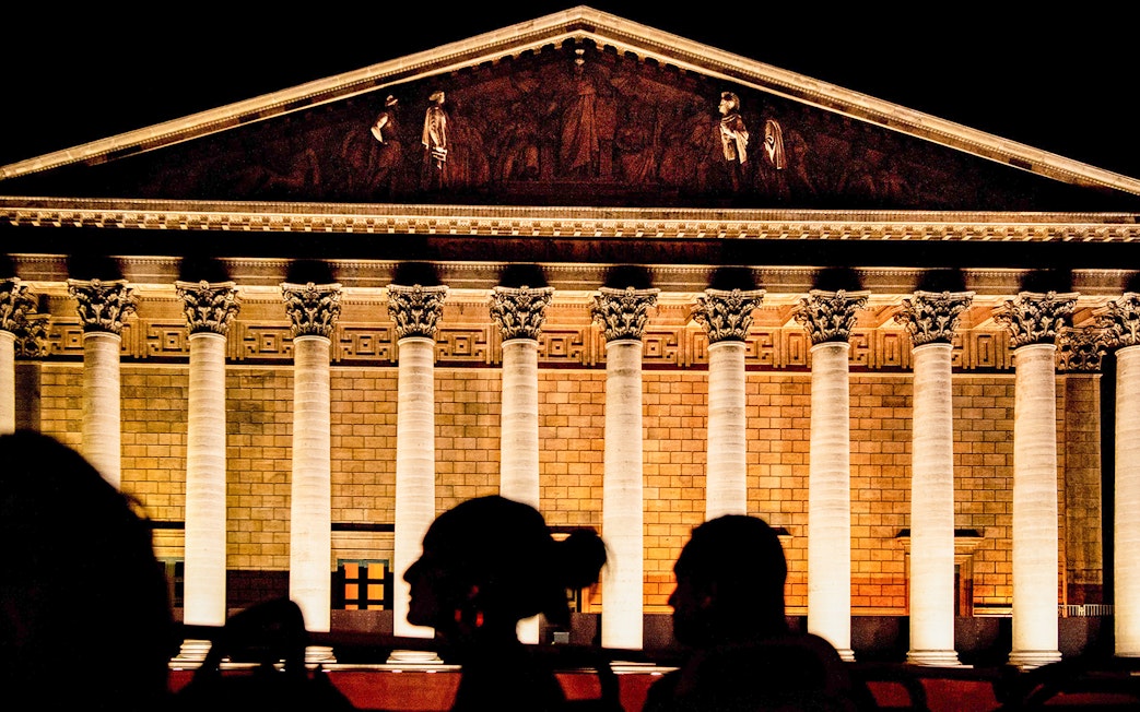 Illuminated facade of La Madeleine in Paris at night with silhouetted figures in foreground.