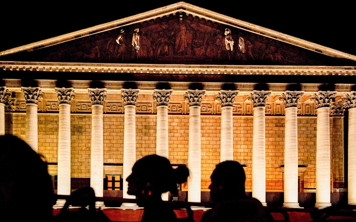 Illuminated facade of La Madeleine in Paris at night with silhouetted figures in foreground.