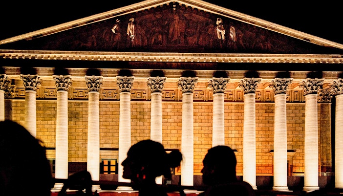 Illuminated facade of La Madeleine in Paris at night with silhouetted figures in foreground.