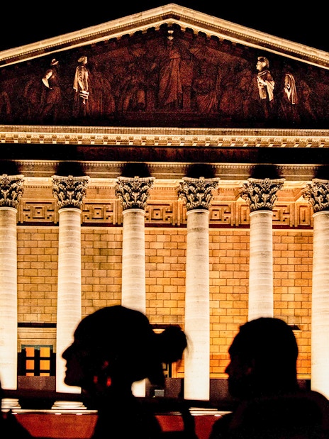 Illuminated facade of La Madeleine in Paris at night with silhouetted figures in foreground.
