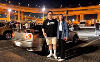 Guests at JDM Tokyo car meetup night tour with skyline and cars in background.