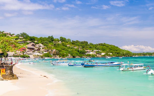 Boats anchored near a sandy beach on Nusa Lembongan, Bali, with lush greenery in the background.