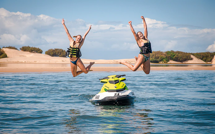 Jet ski with two people jumping into the water, beach in the background.