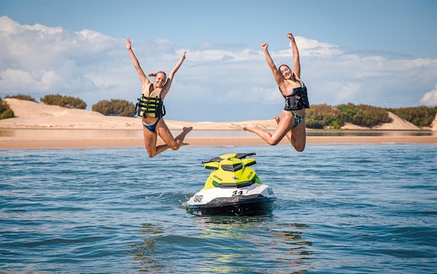 Jet ski with two people jumping into the water, beach in the background.