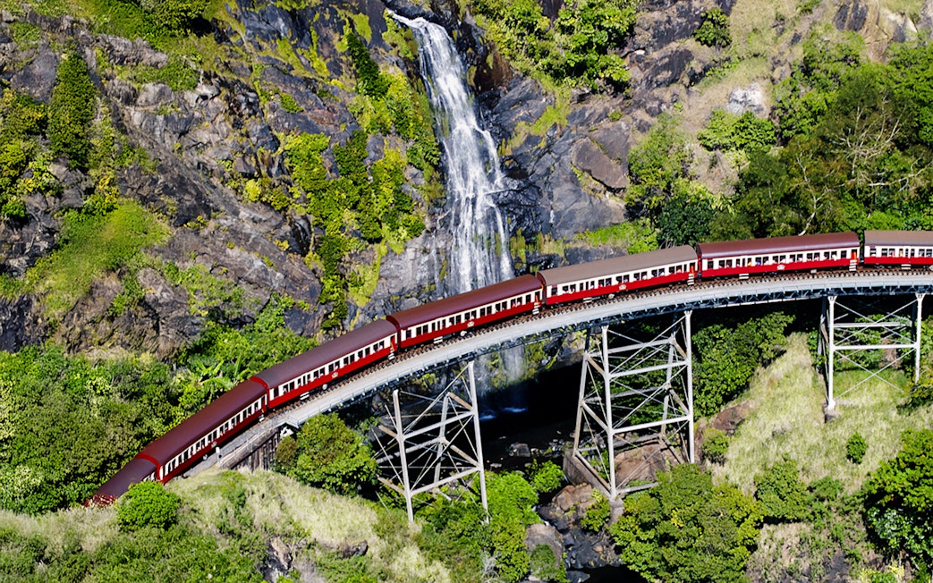 Aerial view of Kuranda Scenic Railway crossing a bridge with a waterfall in the background.