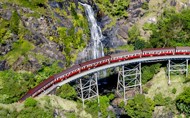 Aerial view of Kuranda Scenic Railway crossing a bridge with a waterfall in the background.