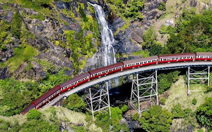 Aerial view of Kuranda Scenic Railway crossing a bridge with a waterfall in the background.